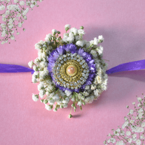 Eco-friendly floral Rakhi with gold and crystal centerpiece, purple asters, white baby's breath, and purple ribbon