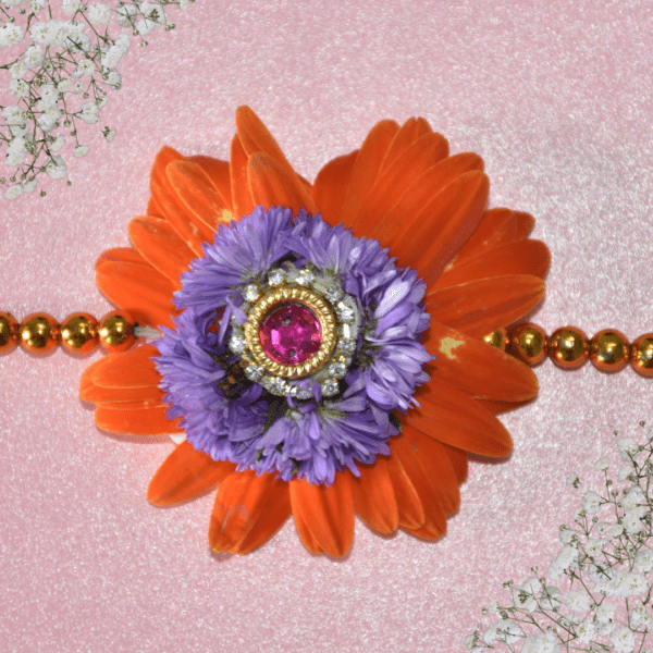 Eco-friendly rakhi featuring orange and purple flowers with pink gemstone centerpiece on golden bead strand