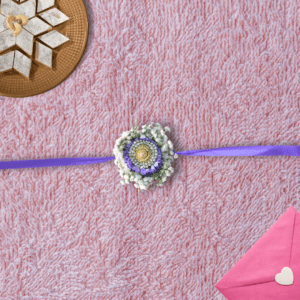 Eco-friendly floral Rakhi with gold and crystal centerpiece, purple asters, white baby's breath, and purple ribbon