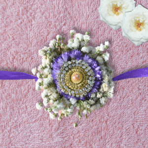 Eco-friendly floral Rakhi with gold and crystal centerpiece, purple asters, white baby's breath, and purple ribbon