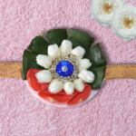 Eco-friendly rakhi featuring white jasmine buds, red rose petals, green leaves, and blue gemstone centerpiece on golden ribbon