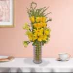 A bunch of yellow roses, yellow carnation, yellow gerbera, and green leaves beautifully arranged in a vase