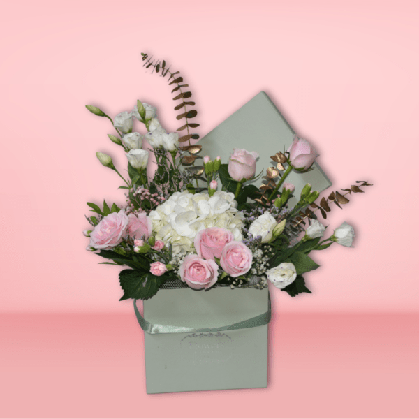 Elegant pastel flower bouquet featuring pink roses, white hydrangeas, and baby's breath in a mint-green envelope-style basket, set against a pink background.