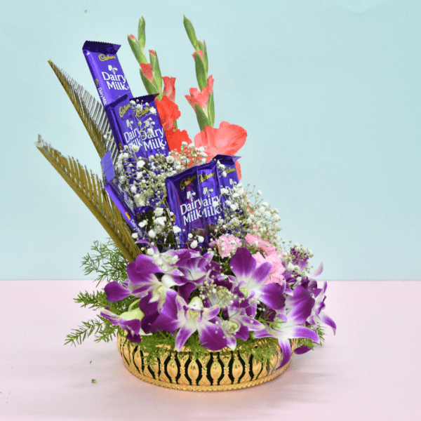 Luxurious flower bouquet with purple orchids, pink carnations, coral gladioli, and Cadbury Dairy Milk chocolates in a golden basket