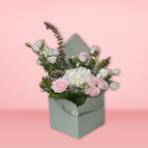 Elegant pastel flower bouquet featuring pink roses, white hydrangeas, and baby's breath in a mint-green envelope-style basket, set against a pink background.