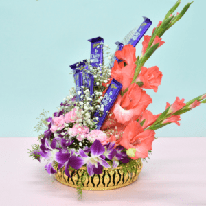 Luxurious flower bouquet with purple orchids, pink carnations, coral gladioli, and Cadbury Dairy Milk chocolates in a golden basket