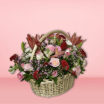 Colorful flower basket arrangement with orange lilies, pink roses, red carnations, and purple flowers in a wicker basket on pink background