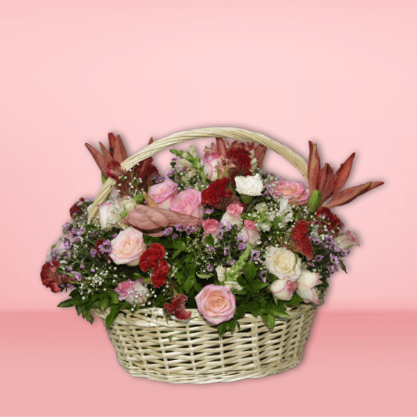 Colorful flower basket arrangement with orange lilies, pink roses, red carnations, and purple flowers in a wicker basket on pink background