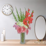 A mixed flower bouquet with red gladiolus, pink carnations, white chrysanthemums, and pink gerbera, arranged in a glass vase.