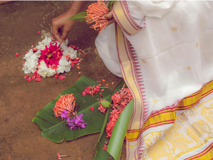 A lady in traditional cotton saree making pookkalam with red, white and purple flowers