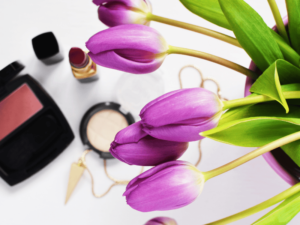 Close-up of purple tulips next to makeup products including lipstick, blush, and compact powder on a white surface