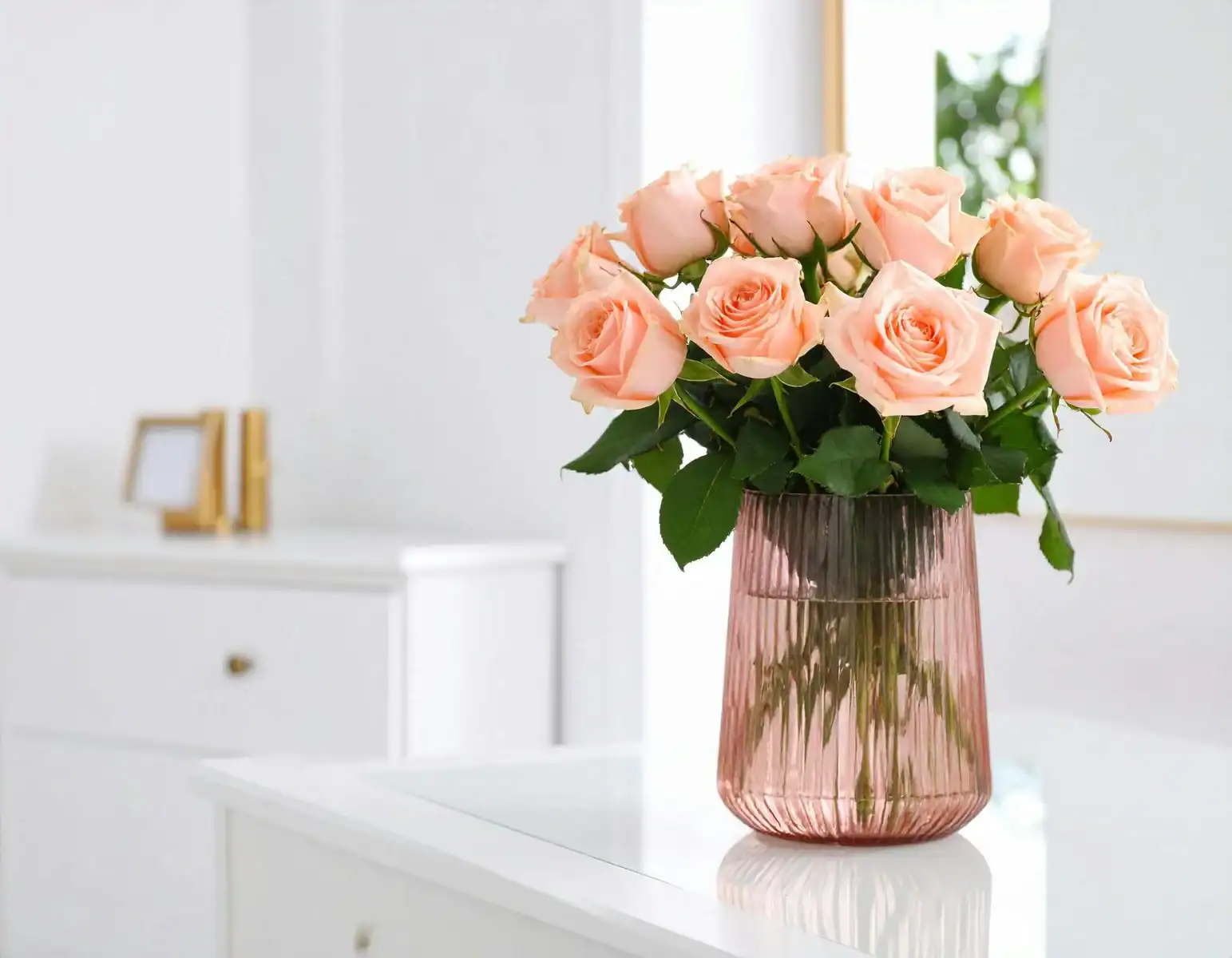 Elegant bouquet of peach roses in a pink fluted glass vase on a white table, minimalist modern home interior decor with a bright room background.