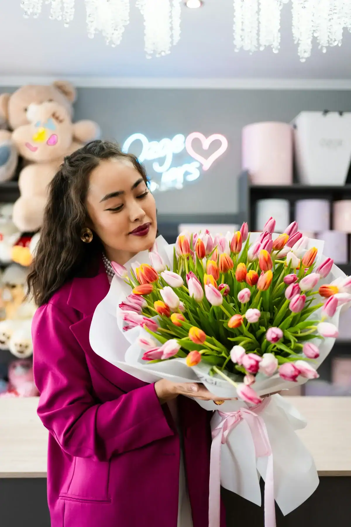 A woman in a magenta blazer holding a large, elegant bouquet of pink, orange, and white tulips wrapped in white paper and a silk ribbon inside a modern flower shop with neon lighting.