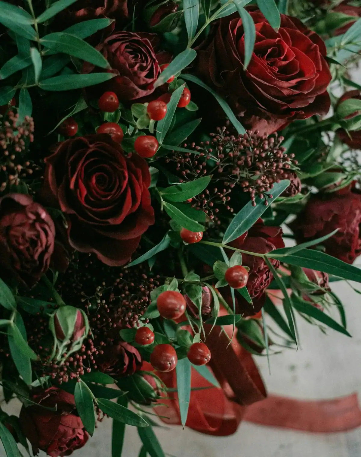 Macro shot of a dark, moody floral arrangement featuring deep red roses, hypericum berries, and various textured green leaves.