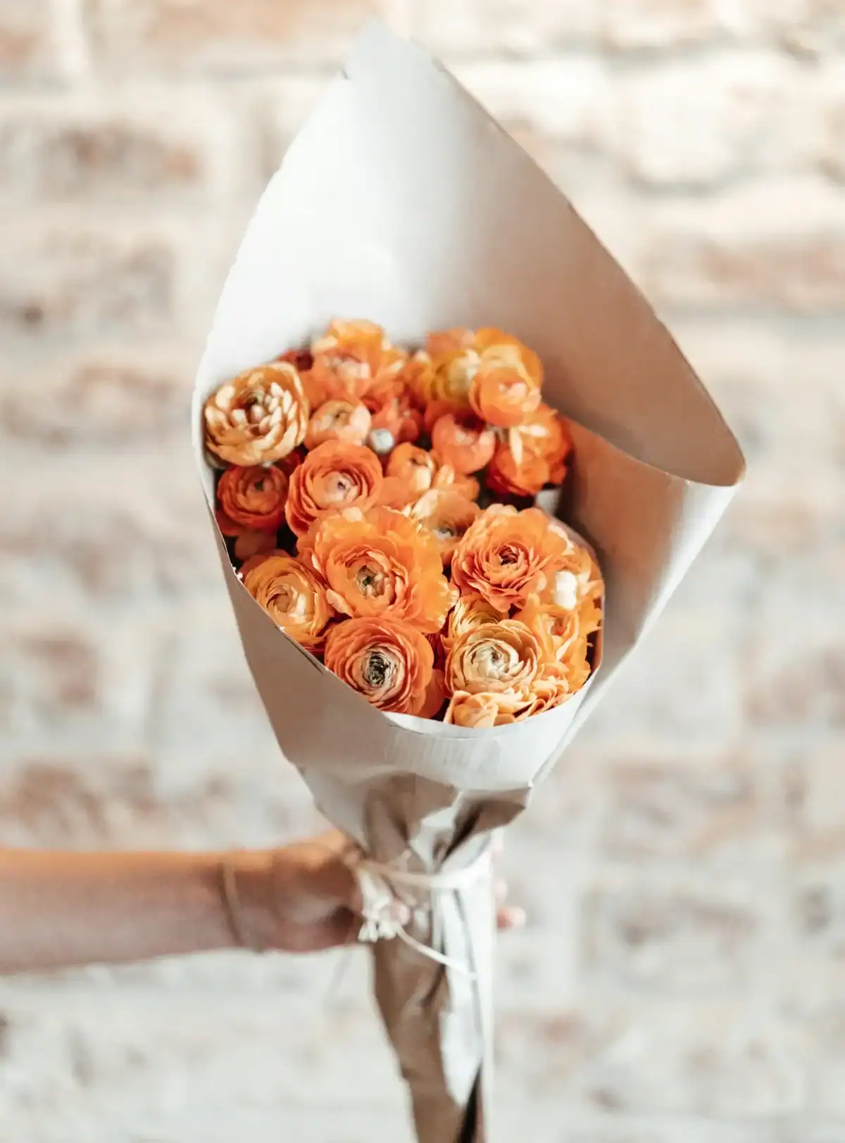A hand holding a rustic bouquet of vibrant orange Ranunculus flowers wrapped in brown kraft paper, set against a blurred neutral brick wall background.