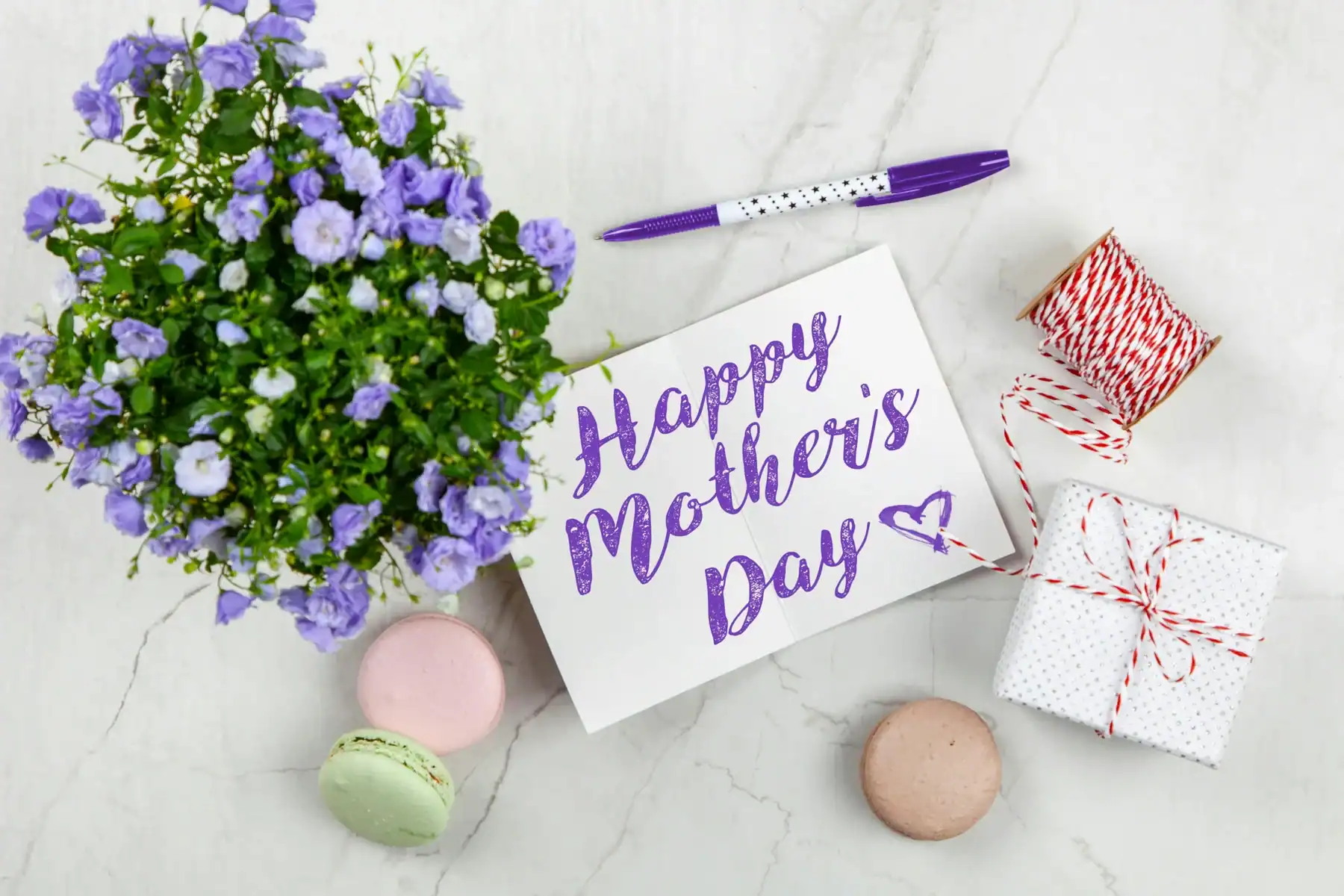 A top-down flat-lay of a "Happy Mother's Day" card with purple calligraphy, accompanied by a potted purple bellflower plant, macarons, a gift box, and a pen on a marble surface.