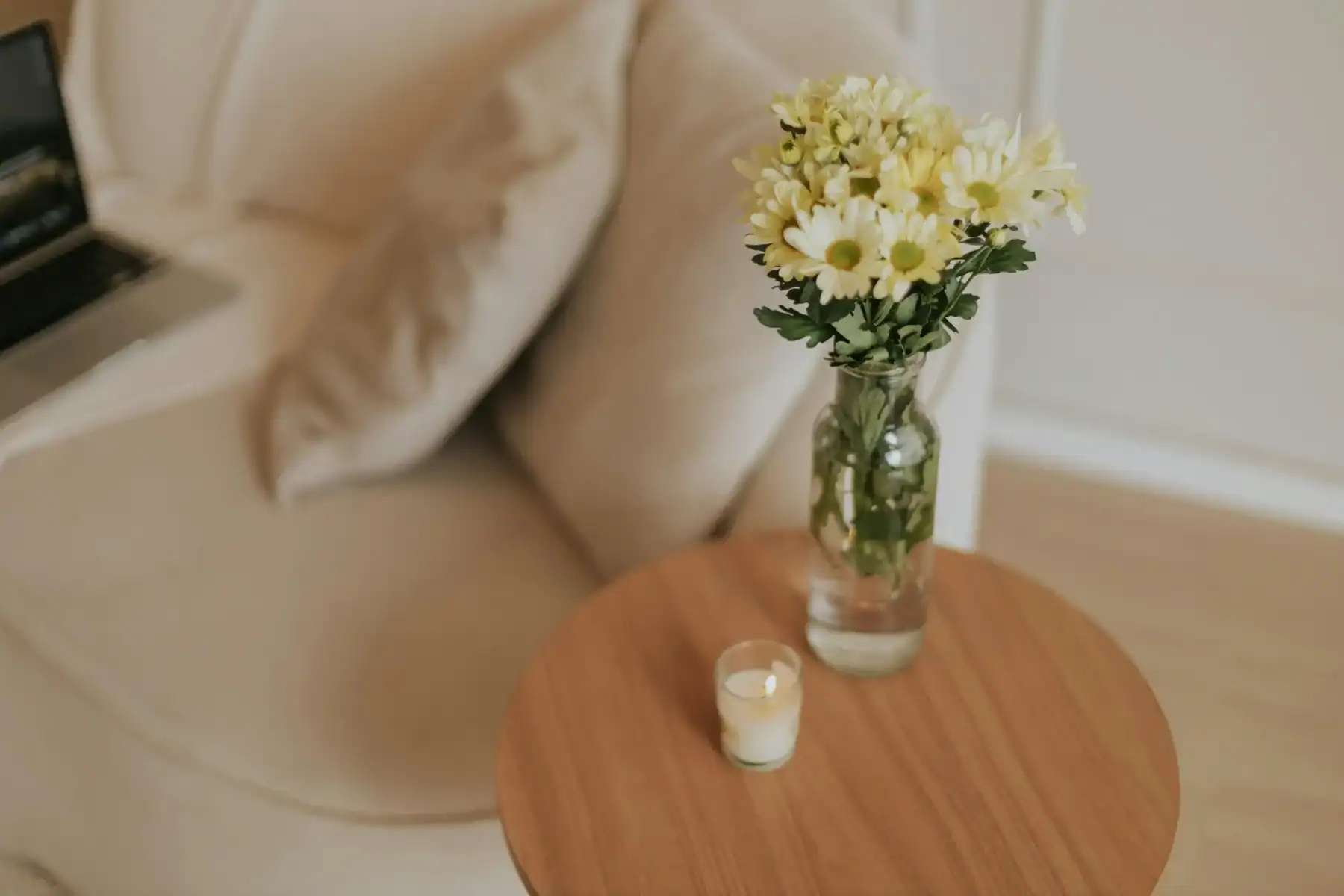 Yellow and white daisy chrysanthemums in a glass bottle vase on a round wooden side table with a lit candle, cozy home floral decor