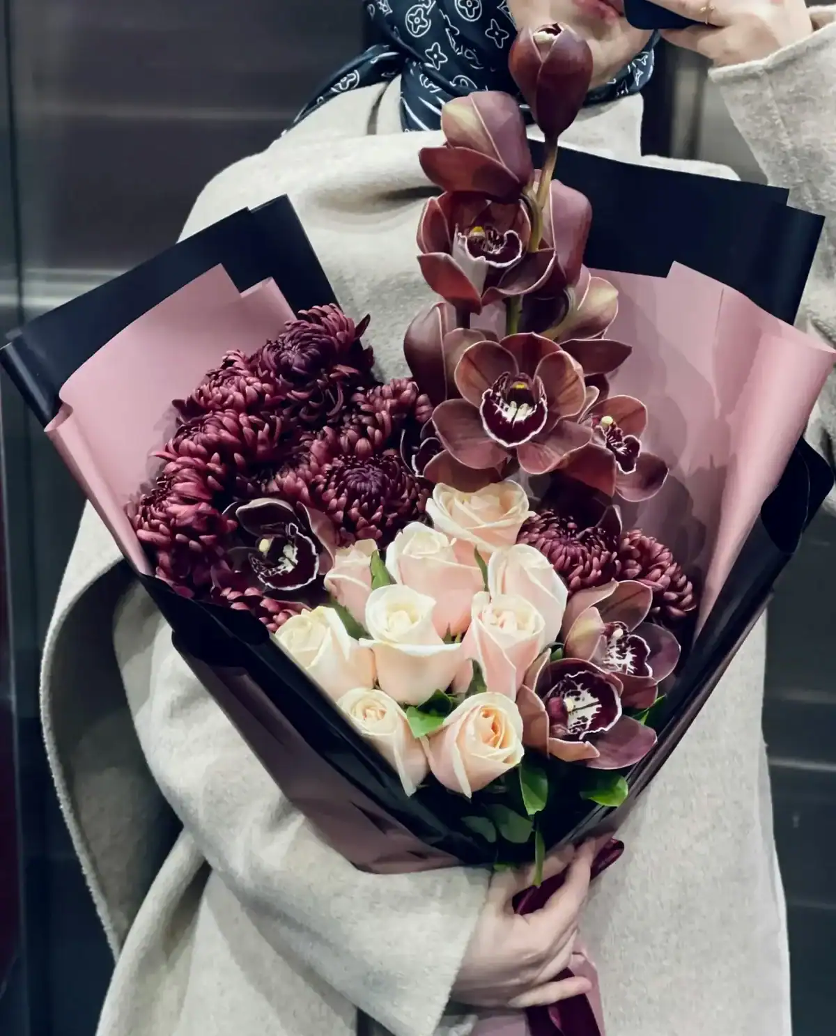 Person holding a dramatic luxury bouquet of burgundy cymbidium orchids, dark maroon spider chrysanthemums and blush pink roses wrapped in black and pink paper, premium flower gift bouquet