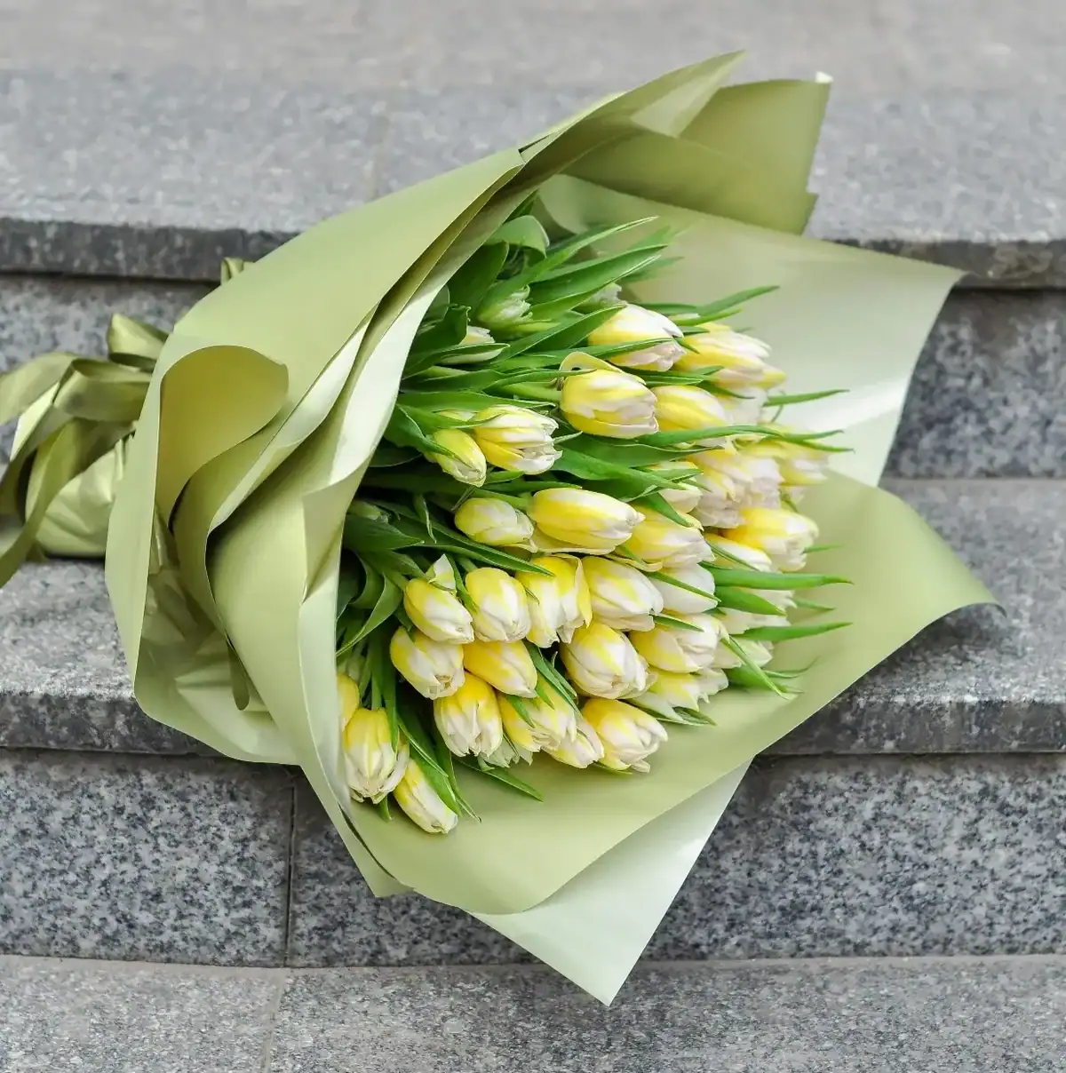 Large bouquet of yellow and white tulips wrapped in sage green paper lying on granite steps, fresh spring flower gift delivery bouquet