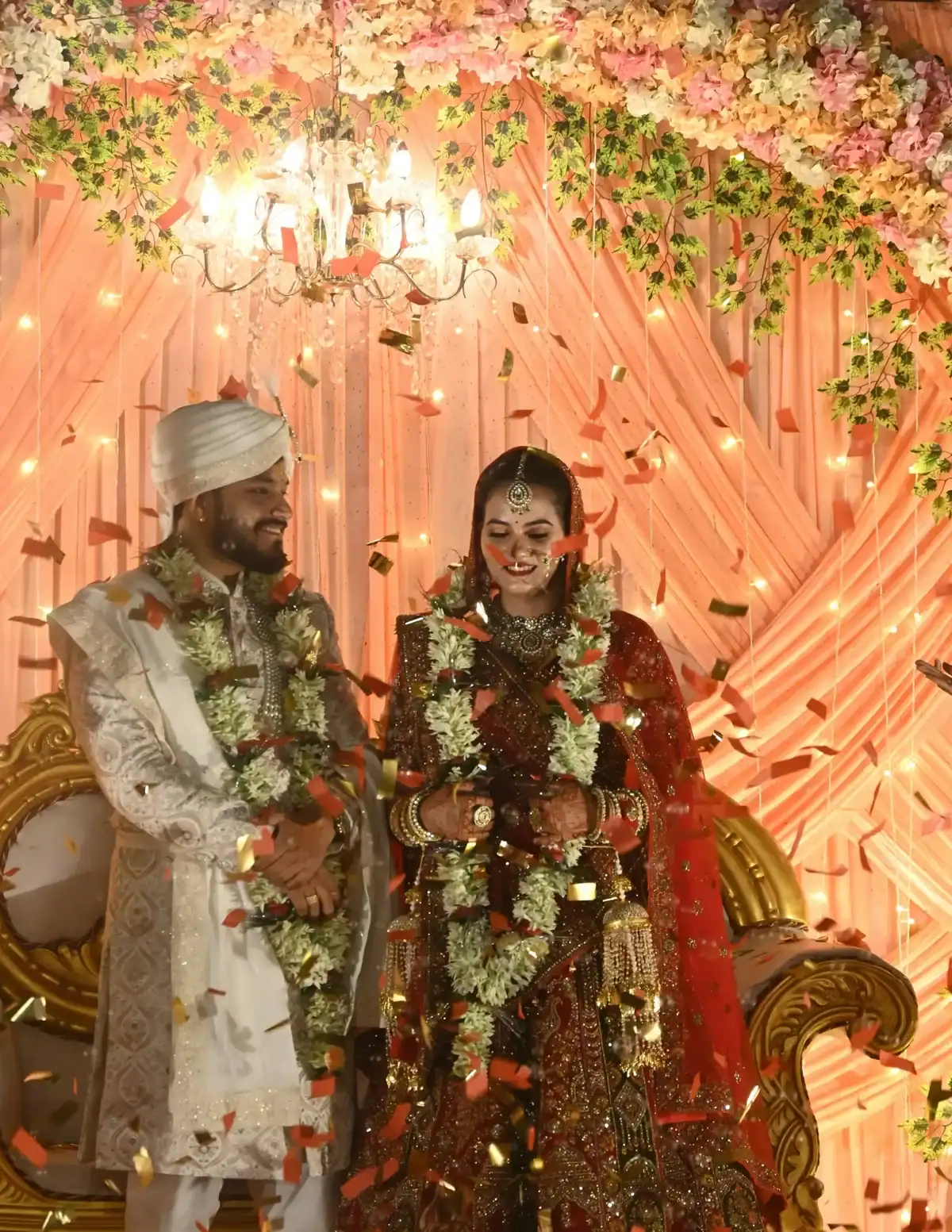 Smiling Indian bride and groom wearing white jasmine varmala garlands under a crystal chandelier with confetti falling, peach floral backdrop Indian wedding ceremony