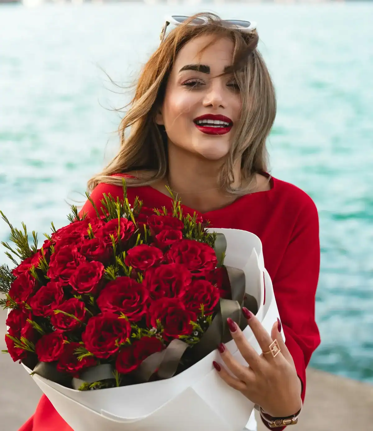 Smiling woman in red dress holding a large red rose bouquet with green fern accents wrapped in white paper near a waterfront, romantic red rose flower gift for girlfriend