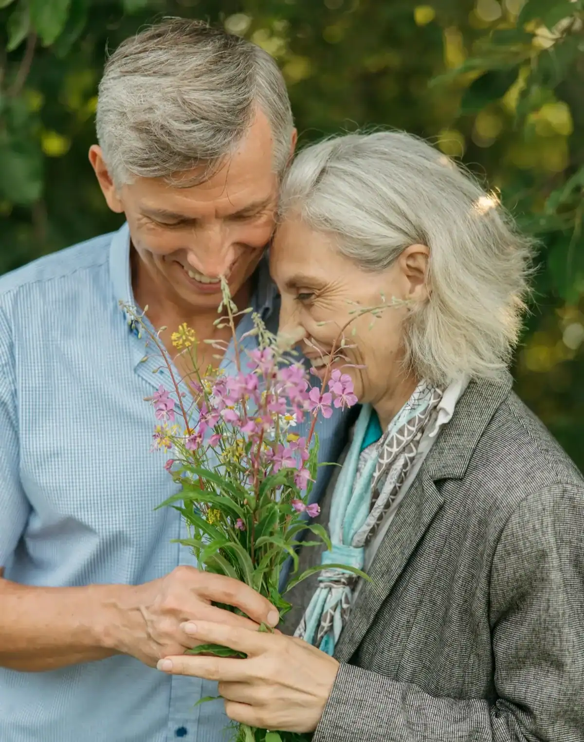Smiling older couple holding and smelling a bunch of pink wildflowers together outdoors in a green garden, romantic flower gift for anniversaries and special occasions