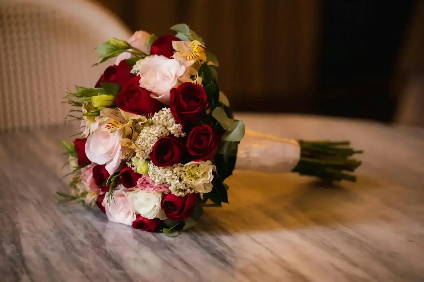 Round bridal bouquet of deep red and blush pink roses with white alstroemeria and green eucalyptus leaves resting on a wooden surface, classic wedding flower bouquet arrangement 