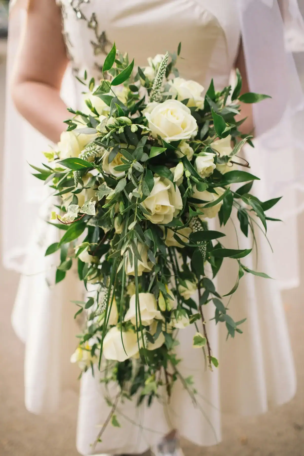 Bride in white dress holding a large cascading bridal bouquet of white roses, ivy, eucalyptus and trailing greenery, elegant waterfall wedding flower arrangement