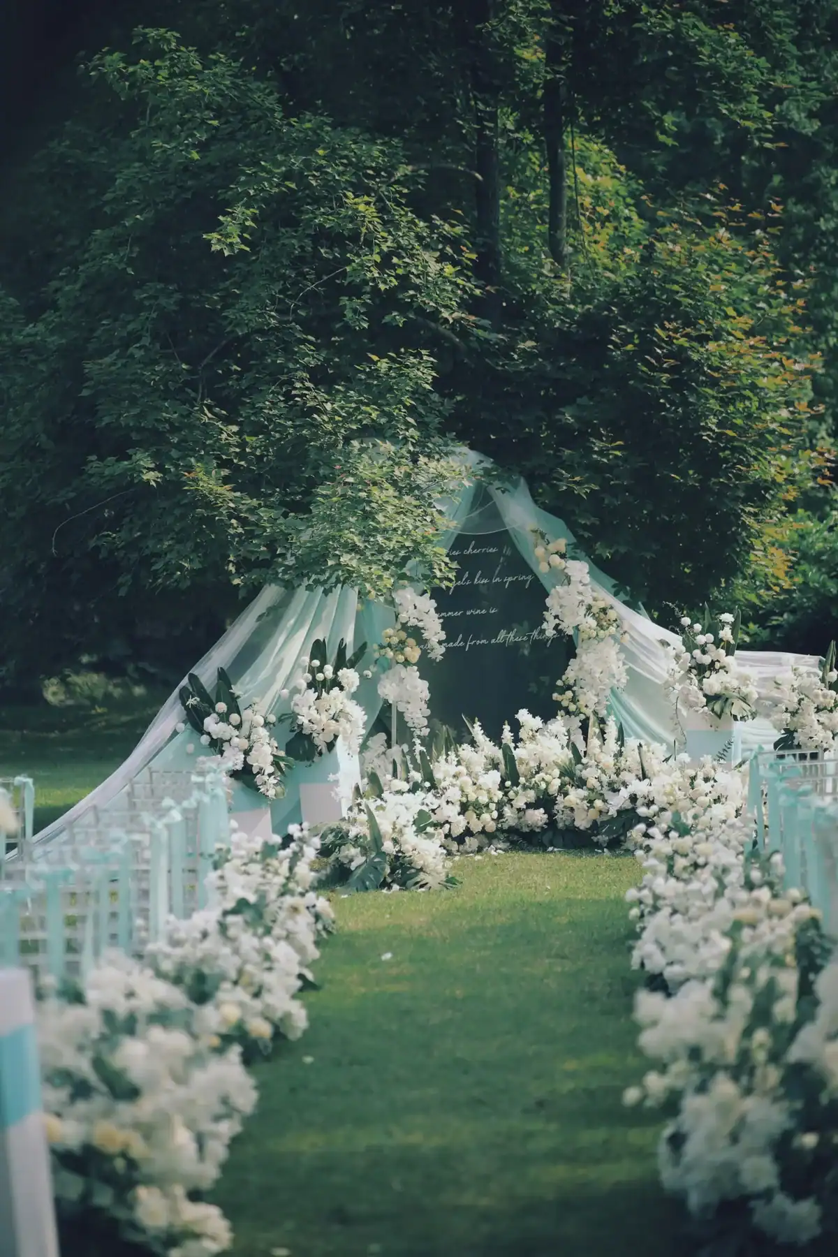 Elegant outdoor garden wedding ceremony aisle lined with white hydrangea and orchid floral arrangements leading to a white flower arch with sheer draping, luxury wedding flower decor
