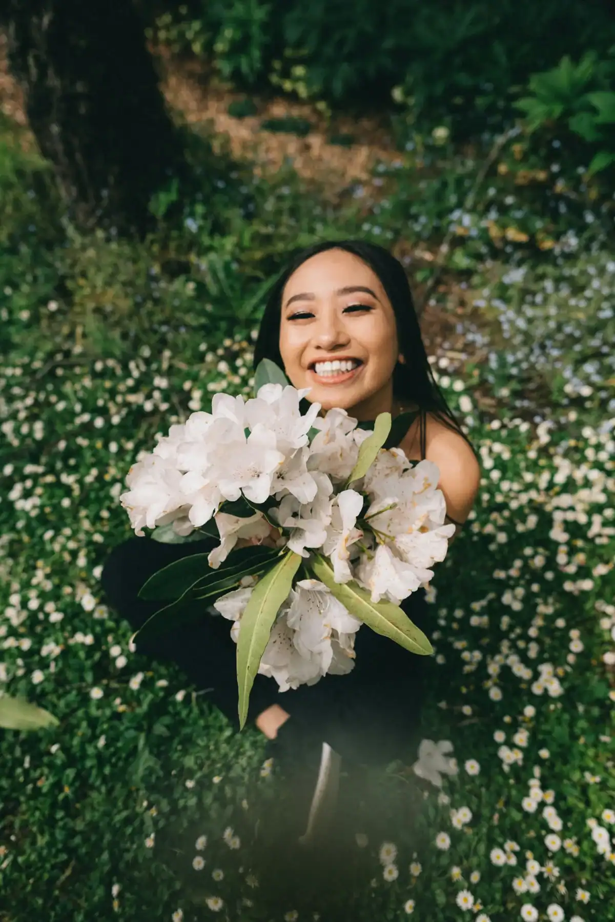Happy young woman lying in a wildflower meadow holding a large bunch of white rhododendron flowers toward camera, joyful fresh flower gift outdoors