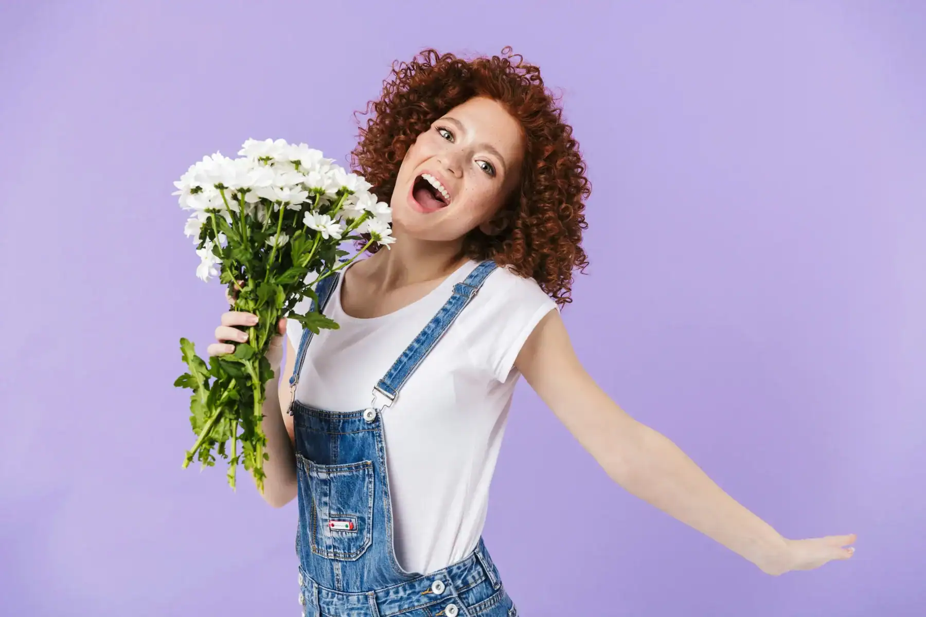 Cheerful curly-haired woman in denim overalls joyfully holding a fresh white chrysanthemum flower bouquet against a purple background, happy flower gift delivery reaction