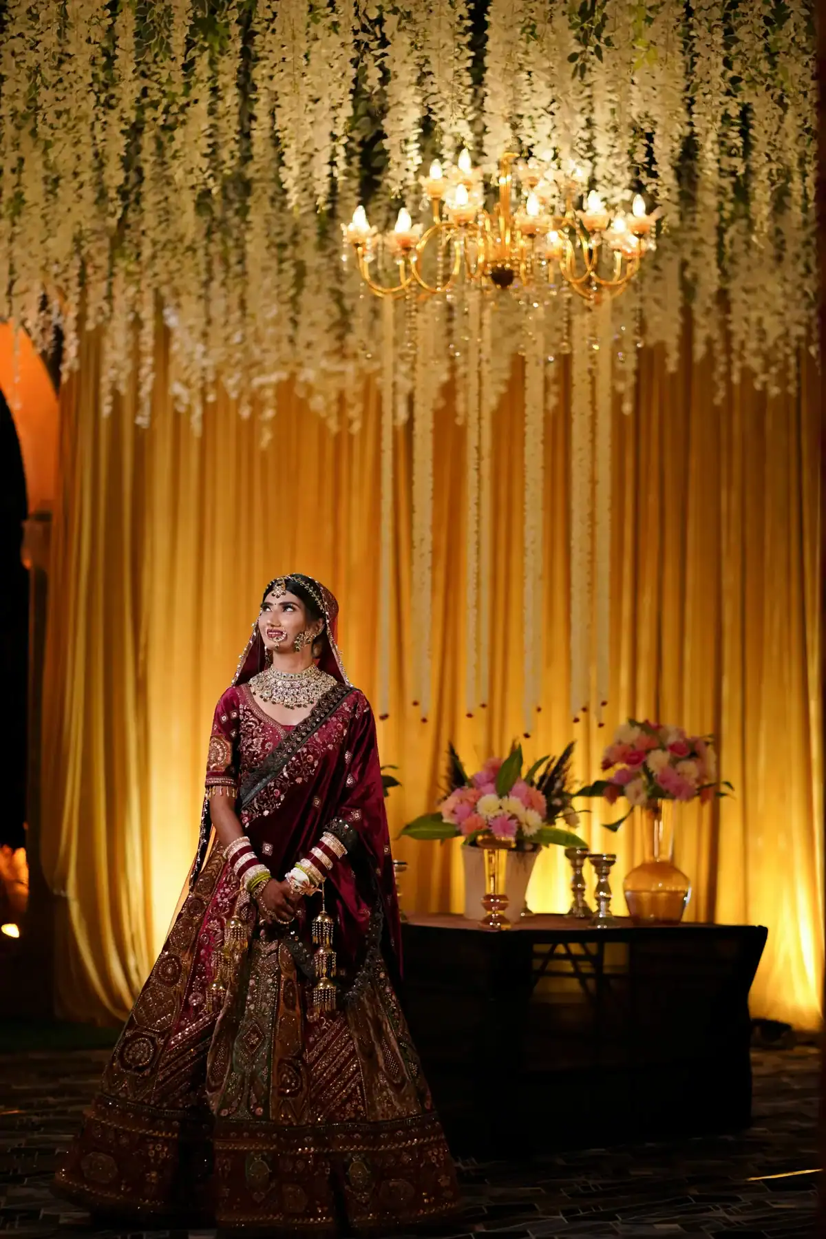 Indian bride in maroon and gold embroidered lehenga standing under a luxurious hanging white orchid and wisteria floral ceiling with gold chandelier, grand Indian wedding flower decoration