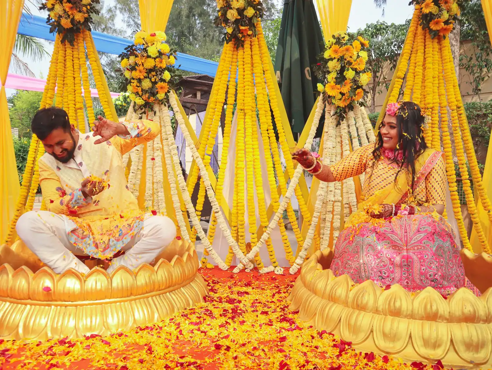 Indian bride and groom celebrating haldi ceremony seated on gold lotus pedestals surrounded by yellow marigold garland mandap decoration and rose petal floor, traditional Indian wedding flower decor 