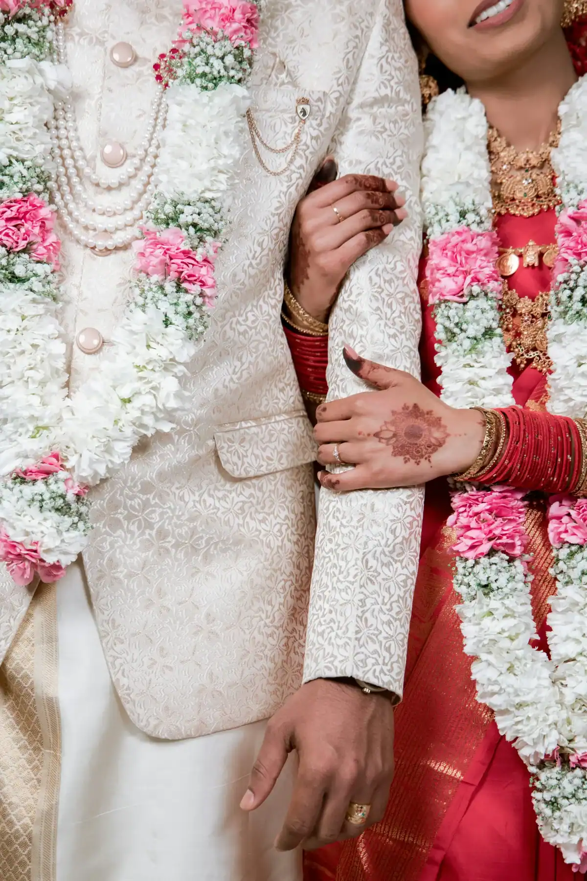 Close-up of Indian bride and groom wearing pink carnation and white baby's breath varmala garlands, bride showing mehndi hands and red bangles in traditional Indian wedding 
